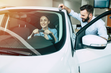 Beautiful young modern couple chooses and buying a new car for the family in the dealership. Woman inside of car tests it