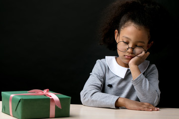 unhappy afro girl is leaning on her chin and looking down while sitting at the table with box gift....