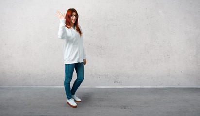 Young redhead girl in an urban white sweatshirt with glasses smiling and saluting and standing on a vintage gray wall