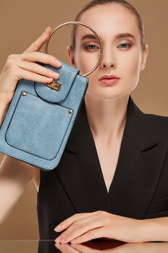 A studio portrait of a chic young lady sporting slicked back hair and looking at the camera through the metal o-ring handle of her blue leather purse. The girl posing over the solid brown background.