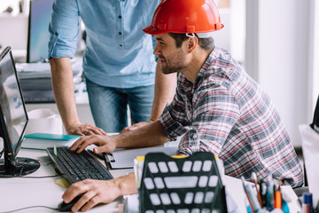 office worker wearing red hardhat is reviewing the plans on his computer. closeup side view photo....