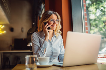 Portrait of young business woman with glasses sitting in cafe in front of her laptop and talking on mobile phone - Powered by Adobe