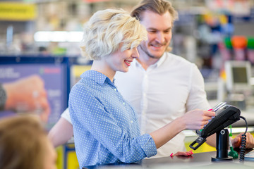 Smiling young blonde lady standing in supermarket shop near cashier's desk paying with credit card,...