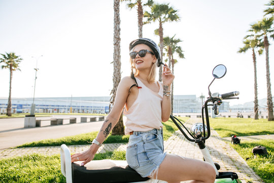 Young caucasian girl wearing helmet and protective glasses sits near electric bicycle on palm alley, blue sky and wide walkway