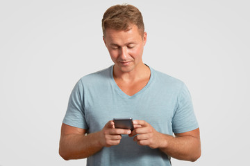 Serious attractive man holds modern smart phone, texts messages, checks his email box, connected to wireless internet, dressed in casual t shirt, isolated on white wall. People and technology concept