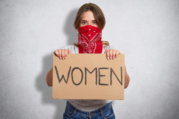 Horizontal shot of beautiful feminist holds plate with inscription Women, belong to special move, has face covered with red badana, isolated over grey concrete wall. Feminism and women concept