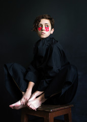 fashion photo girl with short curly hair in black clothes with a high collar with red paint under her eyes sits on a stool