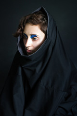 Naklejka premium fashion photo portrait of a girl with curly hair in black clothes with blue paint in front of her eyes in a hood