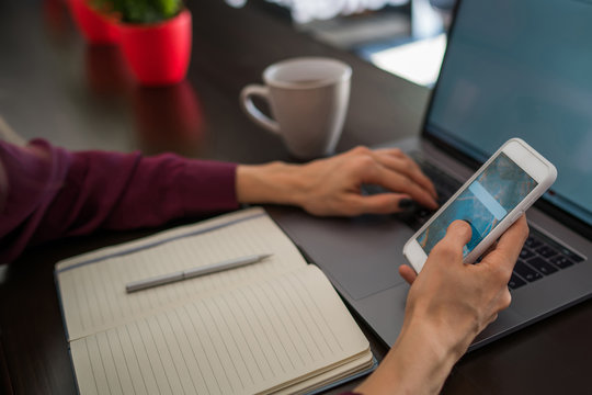 Selective focus on smartphone device which female person hold front open laptop computer sitting at table in cozy interior. Cropped view of woman synchronizing mobile phone and notebook.