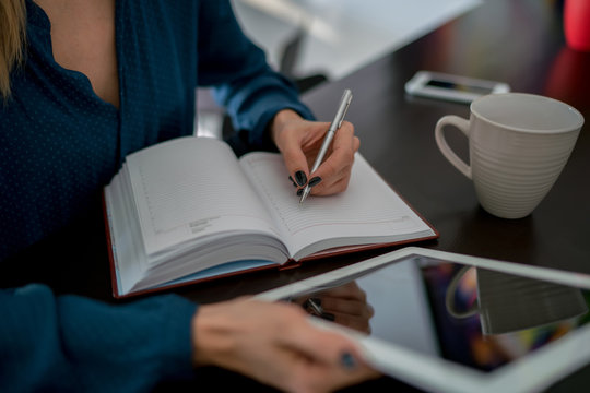 Female author of journalism field writing new article down lined paper pages of notebook. Close up view of women's hands holding white touch pad with refrection on screen. Use of modern technologies