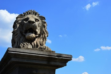 Lying stone lion statue guarding Budapest Chain Bridge, alson known as Szechenyi Landhid, blue summer skies with some clouds in background