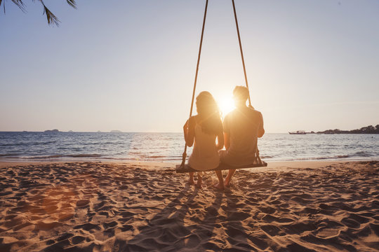 beach holidays for romantic young couple, honeymoon vacations, silhouettes of man and woman sitting together on swing and enjoying sunset