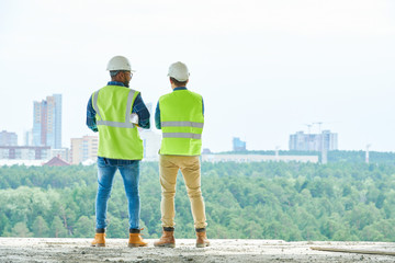 Back view of two men in hardhats and waistcoats standing on construction site and admiring view of...