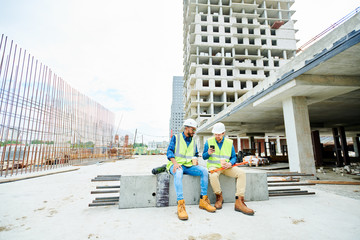 Two men in helmets and waistcoats sitting on concrete beam and browsing smartphone while resting during break on construction site