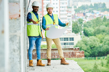 Two men in hardhats and waistcoats holding blueprint while inspecting unfinished building on construction site