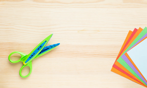 Color paper for origami is a fan, scissors with a wavy edge. On a light wooden table