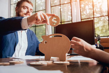 Businessman holds a coin near wooden piggy. Money box. Collecting money with earning bank.