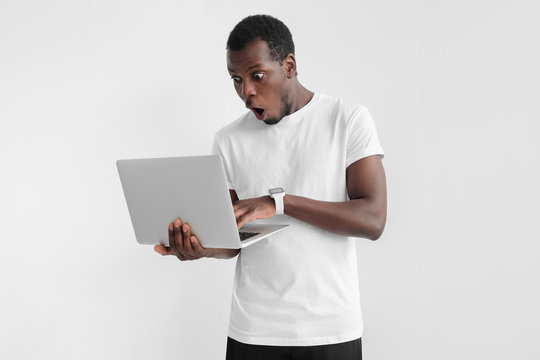 Horizontal picture of young African American man pictured isolated on gray background standing with open laptop in hands, looking at screen with surprised face as if having seen something beneficial