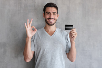 Portrait of young smiling man in t shirt, holding credit card and showing okay sign, standing...