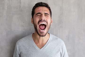 Closeup portrait of screaming with closed eyes crazy young man, standing against gray textured wall