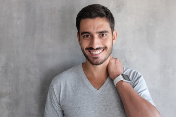 Indoor closeup of young good looking European man standing against gray textured wall, dressed in t-shirt, showing friendly toothy smile,  satisfied and confident