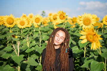 hippie girl with dreadlocks peacefully stands on the field among the sunflowers. A beautiful girl enjoys the warmth of the sun. Summer is outside the city.