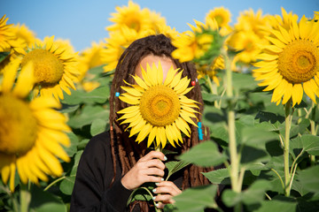 girl with dreadlocks hides her face behind a sunflower. Naughty girl playing hide and seek in the...
