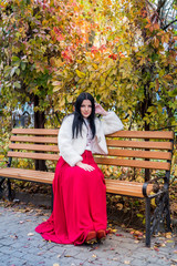 Attractive brunette woman in red dress sitting on bench in autumn park