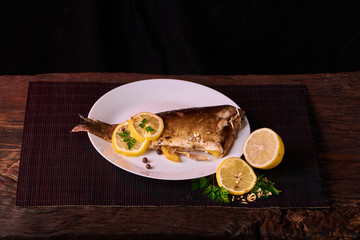 Baked fish with lemon, spices and herbs on white plate on wooden background without head