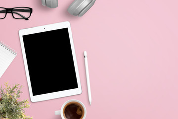 Tablet mockup on pink work desk surrounded with headphones, glasses, notepad, plant, cup of coffee and pen. Free space beside for text. Flat lay composition.