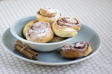 Cinnamon snails called kanelbullar, very tasty sweet rolls in blue bowl on floral pattern tablecloth, raw cinnamon