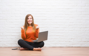 Young adult woman sitting on the floor over white brick wall using computer laptop screaming proud and celebrating victory and success very excited, cheering emotion