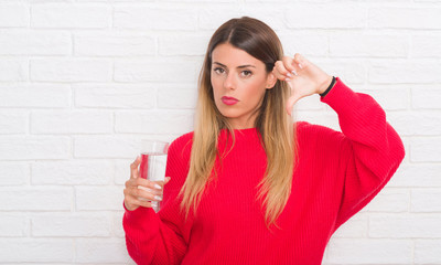 Young adult woman over white brick wall drinking glass of water with angry face, negative sign...