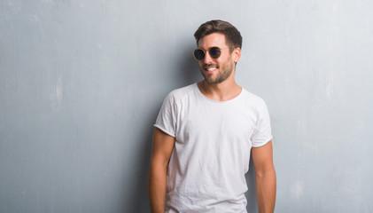 Handsome young man over grey grunge wall wearing sunglasses looking away to side with smile on...