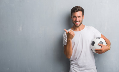 Handsome young man over grey grunge wall holding soccer football ball happy with big smile doing ok...