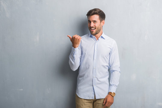 Handsome young business man over grey grunge wall wearing elegant shirt smiling with happy face looking and pointing to the side with thumb up.