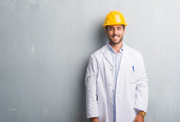 Handsome young engineer man over grey grunge wall wearing safety helmet with a happy face standing and smiling with a confident smile showing teeth
