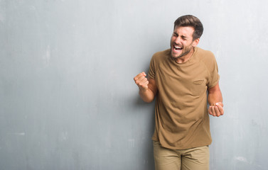 Handsome young man over grey grunge wall very happy and excited doing winner gesture with arms raised, smiling and screaming for success. Celebration concept.