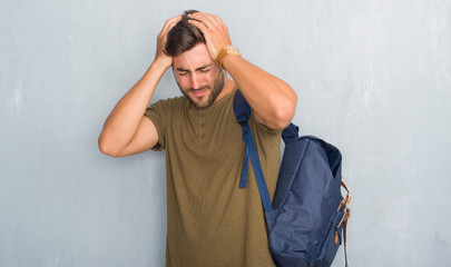 Handsome tourist young man over grey grunge wall wearing backpack suffering from headache desperate...