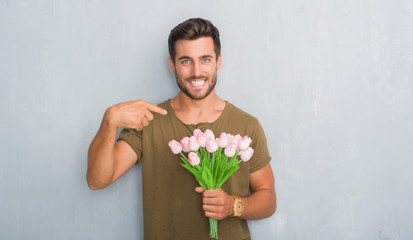 Handsome young man over grey grunge wall holding flowers bouquet with surprise face pointing finger to himself