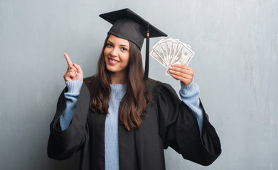 Young brunette woman over grunge grey wall wearing graduate uniform holding dollars surprised with an idea or question pointing finger with happy face, number one