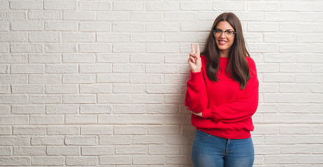 Fototapeta na wymiar Young brunette woman standing over white brick wall showing and pointing up with fingers number two while smiling confident and happy.