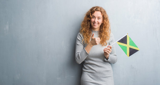 Young redhead woman over grey grunge wall holding flag of Jamaica happy with big smile doing ok sign, thumb up with fingers, excellent sign