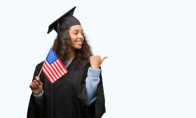 Young hispanic woman wearing graduation uniform holding flag of United States pointing and showing...