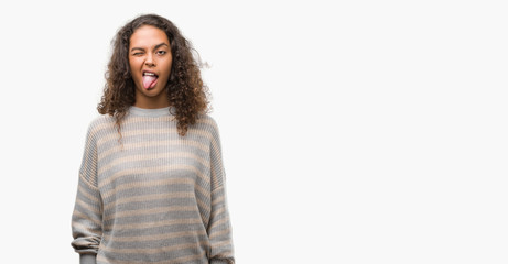 Beautiful young hispanic woman wearing stripes sweater sticking tongue out happy with funny expression. Emotion concept.