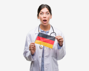 Young hispanic doctor woman holding flag of germany scared in shock with a surprise face, afraid and excited with fear expression