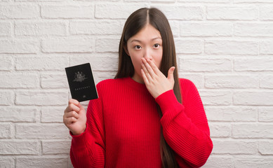 Young Chinese woman over brick wall holding passport of Australia cover mouth with hand shocked with shame for mistake, expression of fear, scared in silence, secret concept