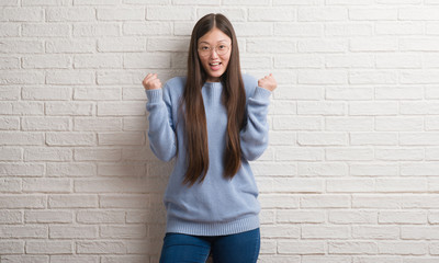Young Chinise woman over white brick wall celebrating surprised and amazed for success with arms raised and open eyes. Winner concept.