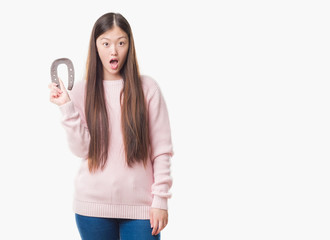 Young Chinese woman over isolated background holding horseshoe talisman scared in shock with a surprise face, afraid and excited with fear expression