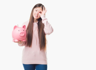 Young Chinese woman over isolated background holding piggy bank with happy face smiling doing ok sign with hand on eye looking through fingers